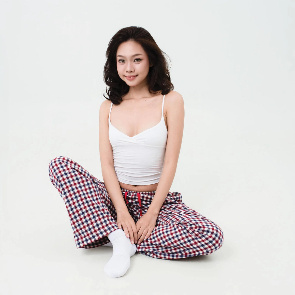 A woman sits cross-legged on a plain white studio background wearing a white ribbed camisole top and red, white, and blue check pyjama bottoms with white socks.